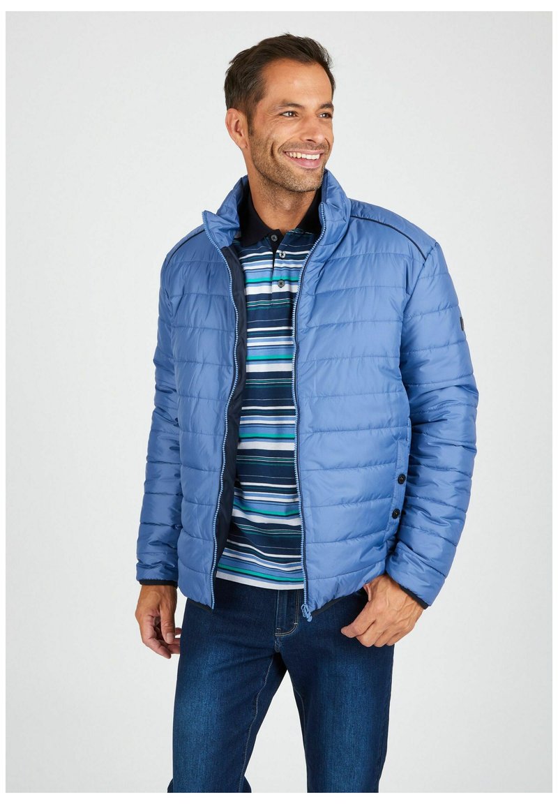 Hellblau gepolsterte Jacke mit Stehkragen, Reißverschluss und schwarzen Akzenten, getragen über einem gestreiften Poloshirt, kombiniert mit dunklen Jeans.
