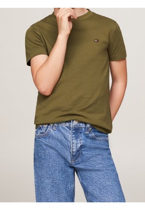 Personne portant un T-shirt vert olive à manches courtes avec un petit logo sur la poitrine et un jean en denim bleu, main reposant près du visage.