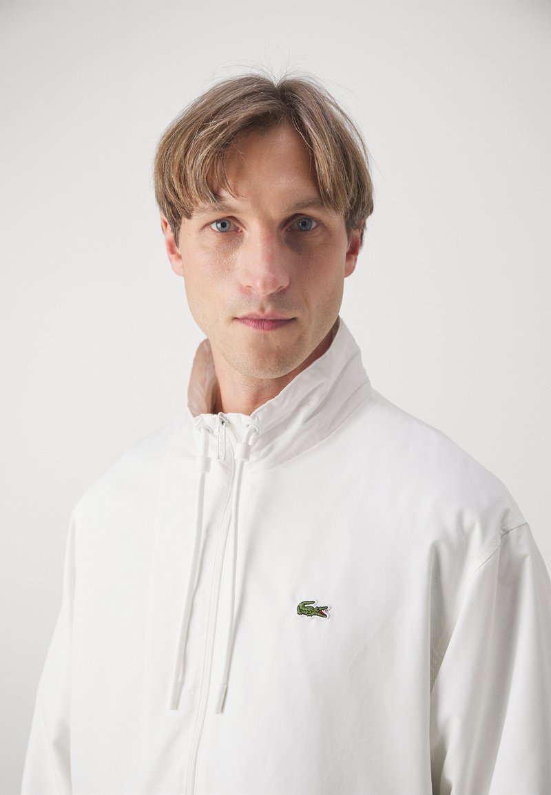 Weiße leichte Jacke mit hohem Kragen, Frontreißverschluss und elastischen Kordelzügen. Besitzt ein kleines grünes gesticktes Logo auf der Brust.