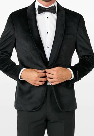 Mężczyzna poprawiający guzik na czarnym aksamitnym smokingu z białą koszulą i czarną muszką.