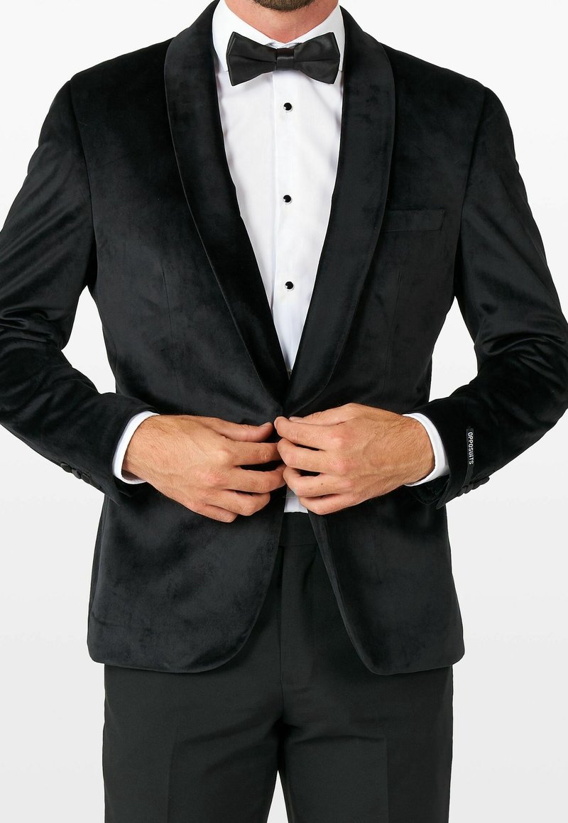 Homme ajustant un bouton sur une veste de smoking en velours noir portée sur une chemise blanche avec un nœud papillon noir.