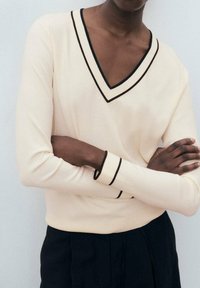 Maglione crema a maniche lunghe con scollatura a V; presenta un bordo scuro a contrasto lungo il colletto e i polsini, con una texture a maglia liscia.