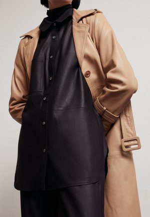 Personne portant une chemise en cuir noire boutonnée sous un trench coat en cuir beige avec ceinture, debout devant un fond clair uni.