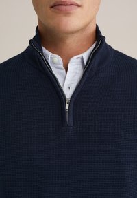 Teksturirani mornarski džemper s poluzip ovratnikom, nošen preko svijetloplave košulje s ovratnikom koja ima neprimjetan prugasti uzorak.