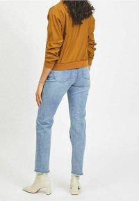 Hnědá bomber bunda s ribovanými manžetami přes světle modré rovné džíny, nošena s béžovými kotníkovými botami na podpatku. Jednoduchý a ležérní design.