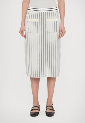 Femme portant une jupe blanche jusqu'aux genoux avec de fines rayures verticales noires, des poches avant, et des chaussures noires à double bride.