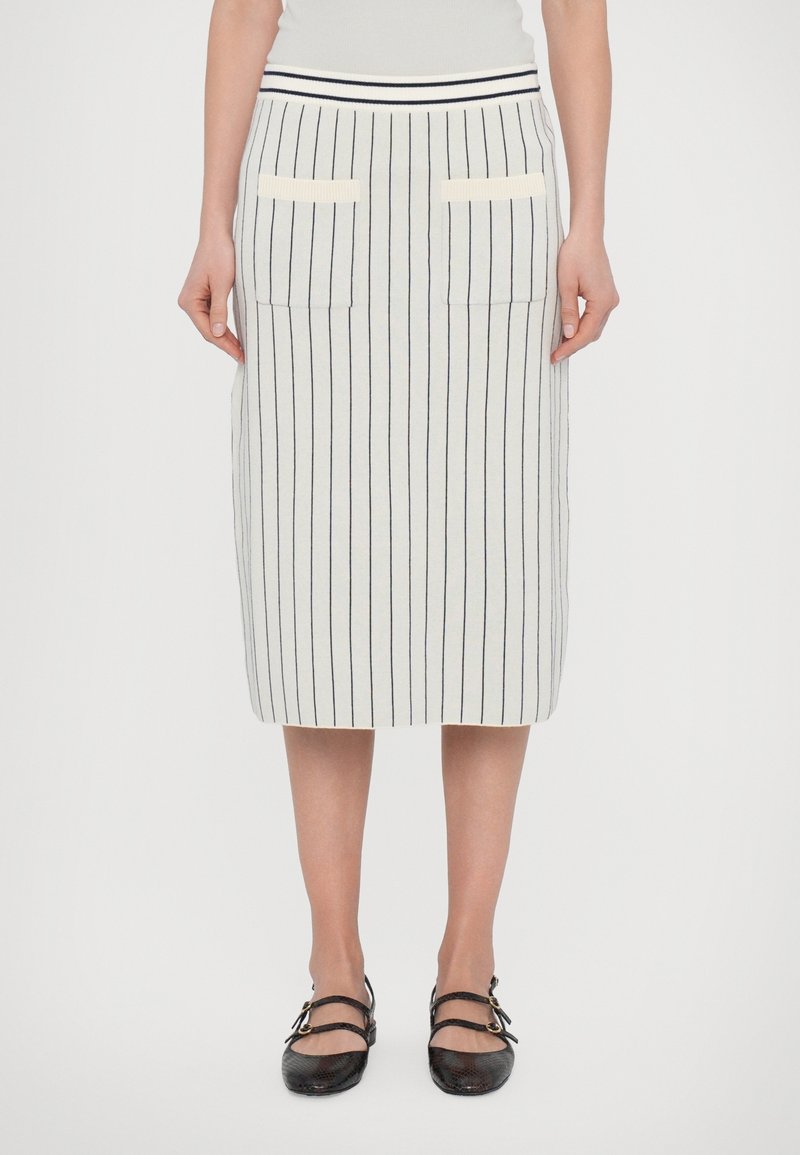 Femme portant une jupe blanche jusqu'aux genoux avec de fines rayures verticales noires, des poches avant, et des chaussures noires à double bride.