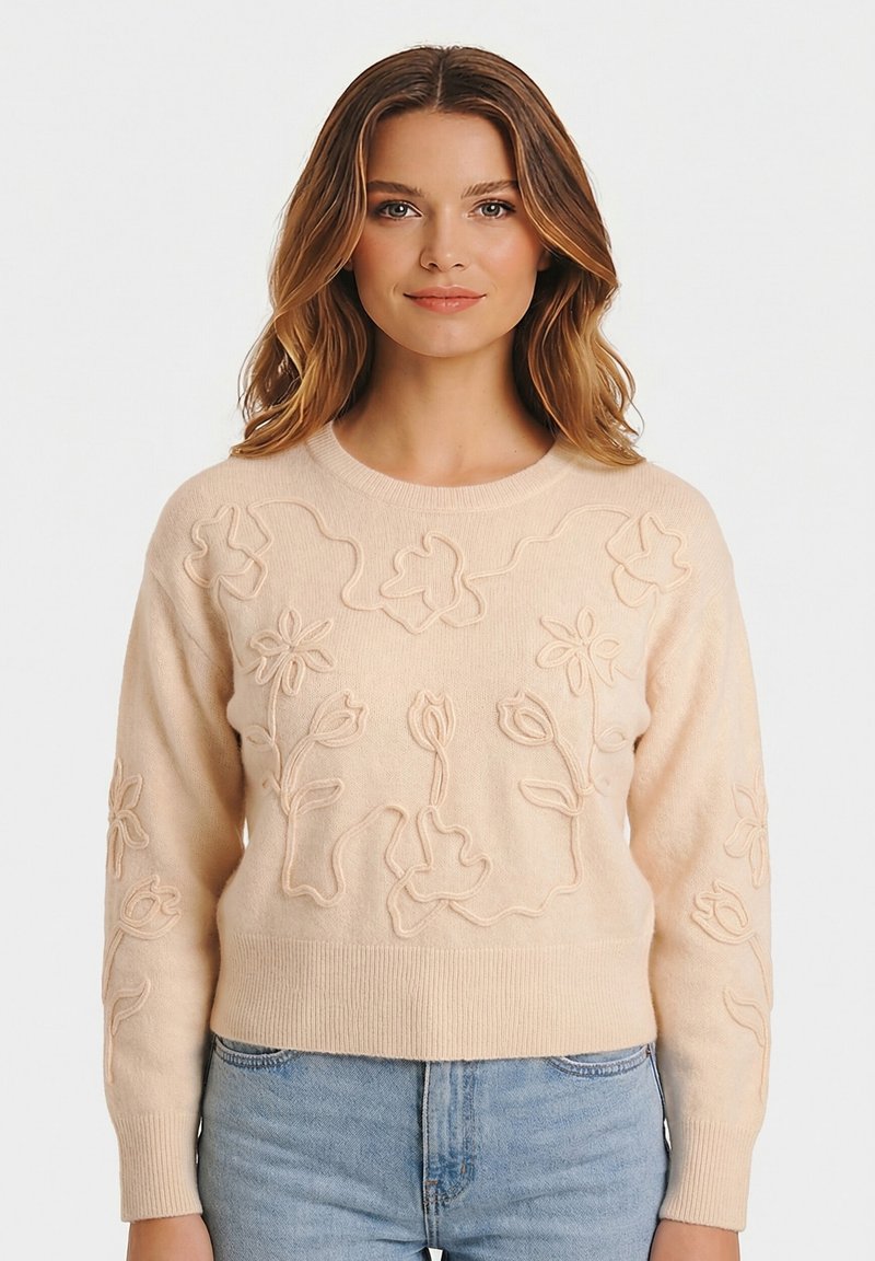 Femme portant un pull beige avec une broderie florale en relief et un jean bleu clair, debout devant un fond blanc uni.