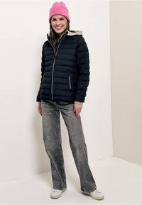 Jonge vrouw die glimlacht, draagt een roze gebreide muts, een zwarte donzen jas met een beige capuchon, grijze wijde jeans en witte sneakers, staand binnenshuis.