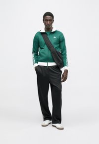 Green zip-up jacket with white collar and three white stripes on sleeves, paired with black trousers and white sneakers, carrying a black bag.