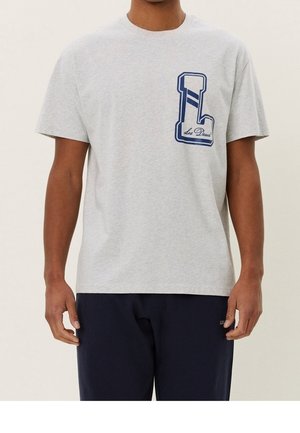 Mężczyzna ubrany w jasnoszary T-shirt z krótkim rękawem, z dużą granatową literą "L" i napisem "Les Deux" na lewej piersi, w połączeniu z ciemnymi spodniami.