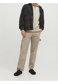 Outfit casual caratterizzato da una camicia button-up a quadri neri e marroni, un maglione zip-up beige, pantaloni beige e scarpe slip-on nere.