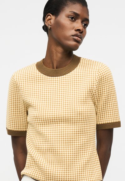 Kvinne med kort hår iført beige og brunt sweaterkortermet genser med hundetannsmønster og ribbestrikket krage og mansjetter, ser litt sidelengs.