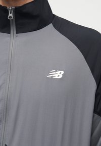 Άτομο που φοράει γκρι και μαύρο τζάκετ New Balance με φερμουάρ και λογότυπο στο αριστερό στήθος, εμφανίζεται από το λαιμό έως τη μέση του κορμού.