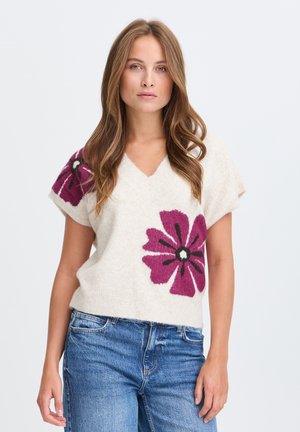 Ung kvinde med langt hår bærer en cremefarvet kortærmet sweater med store lilla blomster og blå jeans, stående mod en hvid baggrund.
