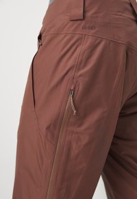 Brune utendørsbukser laget av lett, vannavvisende materiale. Har glidelåslommer og en glatt tekstur.