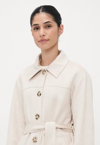 Beige, soft fabric coat with a large collar, five buttons, and a waist tie. Features a smooth texture and subtle stitching details.