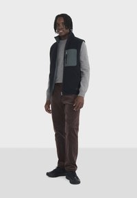 Gilet en polaire noir par-dessus un pull gris, pantalon en velours côtelé brun et baskets noires; présente une poche poitrine zippée et un look globalement texturé.