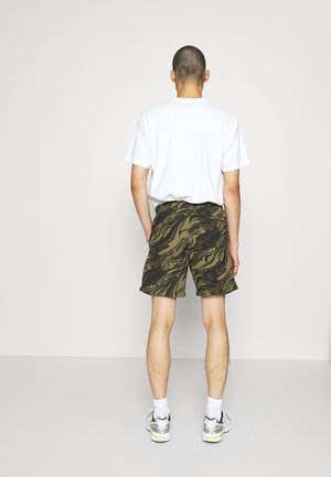 Weißes Baumwoll-T-Shirt für Herren und grüne Camouflage-Shorts mit wirbelnden Mustern. Das Modell trägt weiße Socken und graue Sneaker. Einfaches Design, lockere Passform.