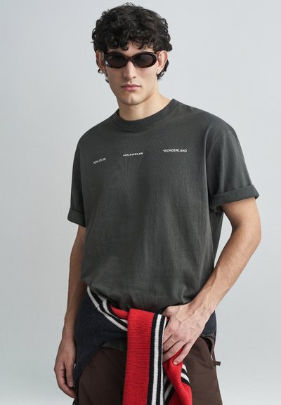 Dunkelgraues T-Shirt mit kurzen Ärmeln und rundem Ausschnitt, mit weißem Text, kombiniert mit einem rot-blauen gestreiften Pullover, der um die Taille gebunden ist.