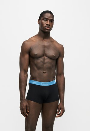 Homme musclé à la peau foncée, debout devant un fond blanc, portant un boxer noir avec une ceinture bleue Calvin Klein.