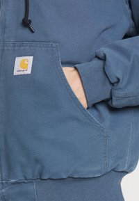 Blauwe hoodie van gestructureerde stof, met een voorzak en een gele Carhartt-logo op een witte patch. Koord bij de hals.