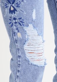 Světle modré džínové kalhoty s vyšívanými květinovými vzory v tmavě modré a krémové barvě. Obsahují poškozené otvory podél stehen a třepené okraje.