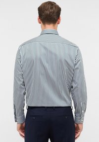 Man gezien van achteren in een verticaal gestreepte langemouwen shirt, ingestopt in donkere marineblauwe broek tegen een effen achtergrond.