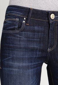Close-up van de tailleband en de voorzak van donkerblauwe denim jeans met oranje stiksel en een zilveren knoopsluiting.