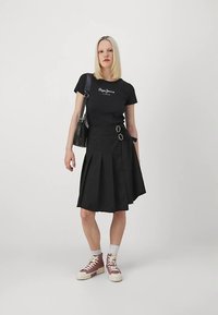 T-shirt preta com logótipo branco, saia preta de cintura subida com pregas e detalhes de fivela, combinada com sapatilhas rosa e brancas e meias curtas brancas.