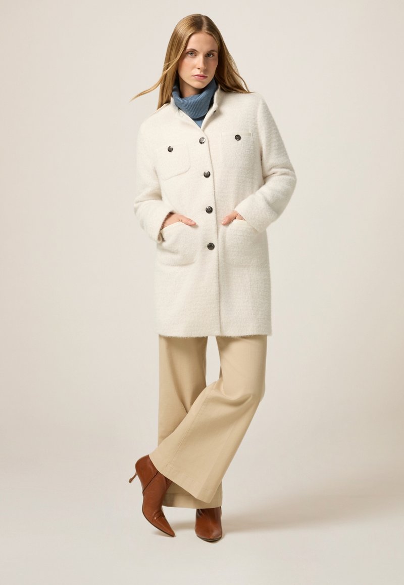 Abrigo de mezcla de lana blanca con un acabado texturizado, dos bolsillos frontales, cierre de botones y un collar, combinado con pantalones de pierna ancha beige y tacones marrones.