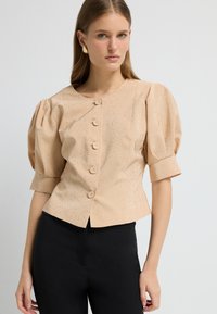 Beige cropped bluse med korte puffærmer, frontknaplukning og subtil spraglet tekstur. Sammen med tætsiddende sorte bukser.