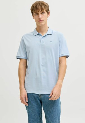 Joven vestido con una camiseta tipo polo de manga corta de color azul claro con un pequeño logo, combinado con vaqueros azules, de pie frente a un fondo sencillo.