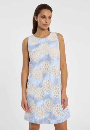 Vrouw in een mouwloze blauw-wit gestreepte jurk met grote witte kanten bloemmotieven, staand tegen een effen achtergrond.