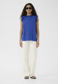 Kék ujjatlan blúz fodros vállrésszel, gombos elejével és laza fazonban, fehér bővérű nadrággal és fekete szandállal párosítva.