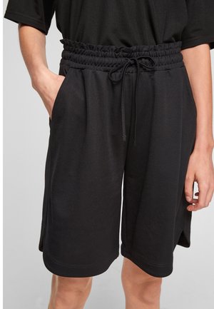 Person wearing black knee-length shorts with elastic waistband and drawstring, standing with one hand in pocket against a light background.