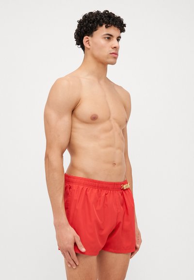 Shorts de bain rouges en tissu léger et lisse, dotés d'une taille élastique et d'un accent décoratif en or à l'avant.