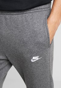 Nahaufnahme einer Person in grauen Nike-Sweatpants mit einer Hand in der Tasche, die das weiße gestickte Nike-Logo am Oberschenkel zeigt.