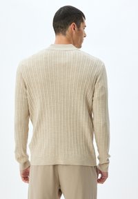 Suéter de punto acanalado beige con mangas largas, que presenta un cuello redondo y un corte holgado. Textura suave y detalle de costura sutil.
