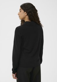 Donna con capelli ricci scuri, che indossa un top nero a maniche lunghe e pantaloni neri, in piedi di profilo contro uno sfondo bianco semplice.