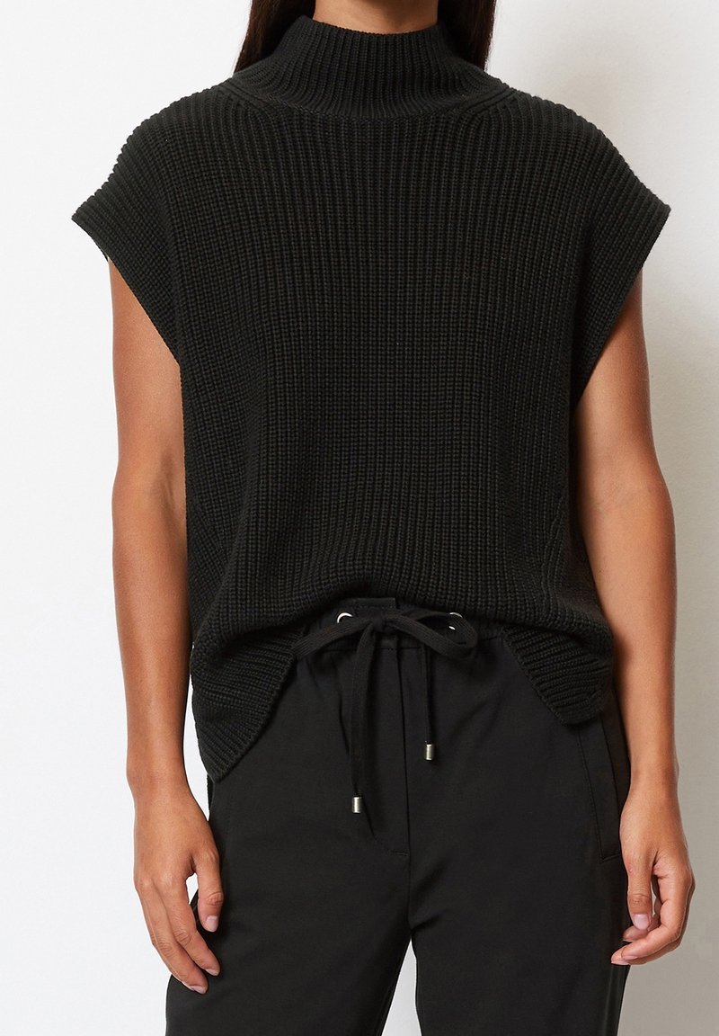 Maglione nero lavorato a maglia con colletto alto, maniche corte e un motivo texture. Abbinato a pantaloni neri con coulisse.
