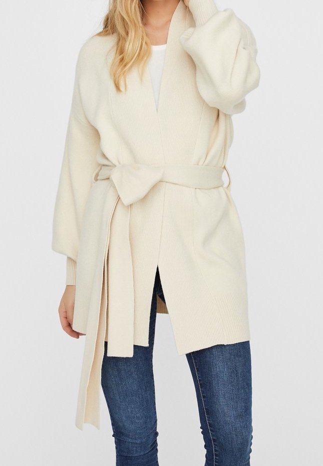 Femme portant un long cardigan crème en tricot avec une large ceinture nouée à la taille, associé à un jean bleu moulant.