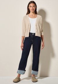 Beige gestrickte Strickjacke mit Dreiviertelärmeln und Knöpfen, kombiniert mit dunkelblauen weit geschnittenen Jeans mit aufgerollten Säumen. Braune Wildleder-Slipper.