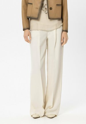 Person trägt beige weite Hosen, beige spitze Schuhe, ein durchscheinendes, geschichtetes Oberteil und eine hellbraune Jacke mit dunklem Besatz.