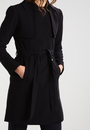 Kvinde iført en sort trenchcoat med bælte, krave, lange ærmer og hænder i forlommerne mod en ensfarvet baggrund.
