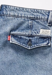 Jeans din denim de culoare albastru deschis prezintă un buzunar aplicat cu un buton negru și un etichetă roșie Levi's. Etichetele includ "Levi Strauss" și "Calitate Garantată."