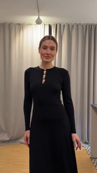 Vrouw draagt een nauwsluitende zwarte jurk met lange mouwen en gouden knopen langs de borst, staand binnenshuis voor beige gordijnen.