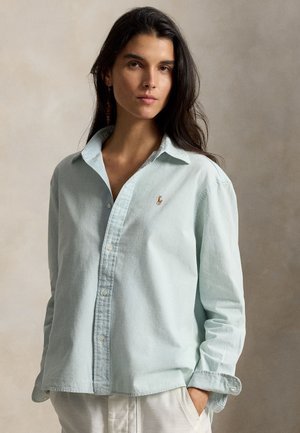 Jeune femme aux longs cheveux foncés porte une chemise bleu clair boutonnée et un pantalon blanc sur un fond beige uni.