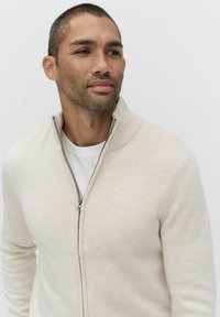 Jasnobeżowy sweter z zamkiem błyskawicznym, z prążkowanym kołnierzem, miękką teksturą i długimi rękawami. Noszony na białej koszulce z okrągłym dekoltem.