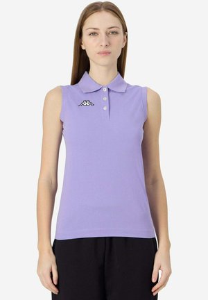 Donna che indossa una polo lavanda senza maniche con tre bottoni e un piccolo logo bianco e nero sul petto, abbinata a pantaloni neri.